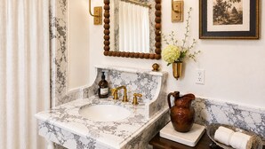 Uma casa de banho com lavatório de mármore, espelho com candeeiros de parede, quadro emoldurado e um vaso com flores.