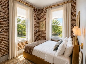 A bedroom with a brick-patterned wall, a bed with white bedding, and a window with curtains. - Neiman's Room at Keystone Hotel - 202 (Savannah)
