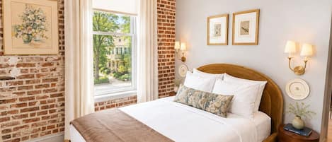 Un dormitorio con pared de ladrillo, un ventanal grande, una cama con ropa blanca y dos lámparas fijadas a la pared.