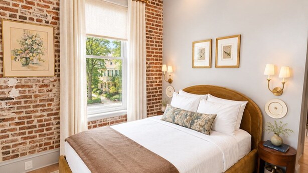 Ein Schlafzimmer mit Ziegelwand, großem Fenster, einem Bett mit weißer Bettwäsche und zwei Wandleuchten.