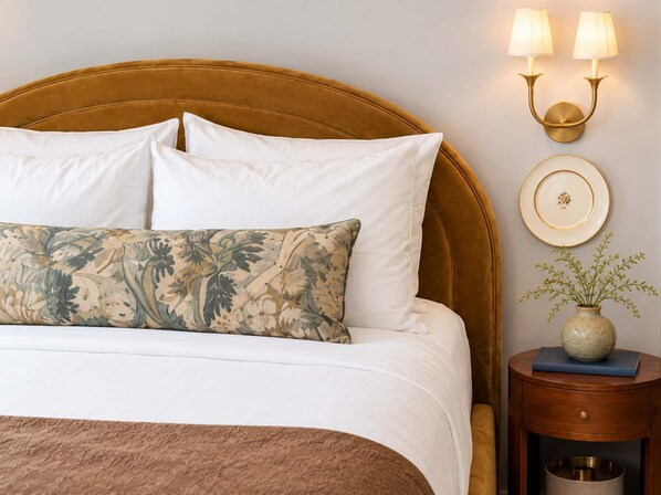 A bed with white bedding, a floral pillow, and a gold-toned headboard. There is a wall-mounted light fixture with two lamps and a decorative plate on the wall.