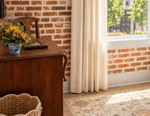 A room with a brick wall, a wooden desk, a vase with flowers, a window with curtains, and a patterned rug.