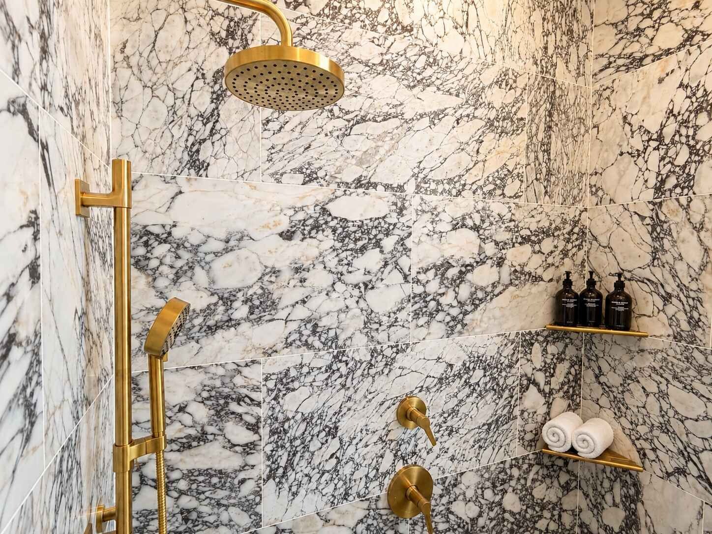 Un baño con ducha de oro, paredes de m ármol y estantes con artículos de aseo.