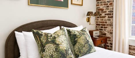A bed with white bedding, two decorative pillows, a nightstand with a lamp, and framed botanical prints on the wall.