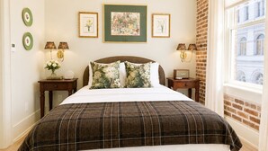 Cama bem arrumada com colcha xadrez, cabeceira e almofadas decorativas. Há quadros nas paredes, uma janela com vista e duas mesas de cabeceira com abajures.