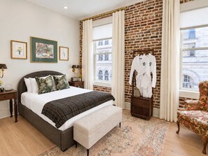 A hotel room with a large bed, a bench, a chair, a brick wall, and a window with a view of buildings. - Grandma's Room at Keystone | Downtown Savannah (Savannah)