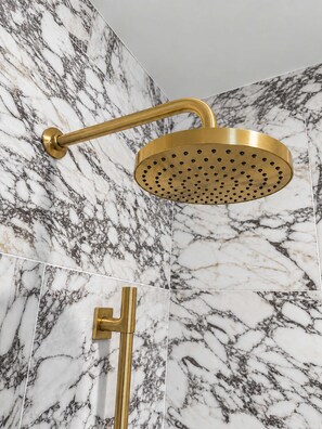 A gold showerhead with a circular design and multiple holes, mounted on a wall with a marble pattern.