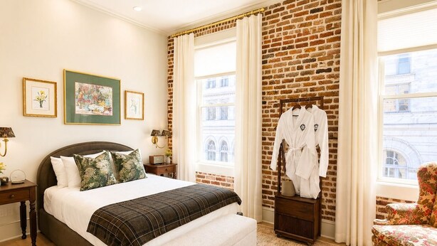 Quarto com parede de tijolos, cama com um banco, uma cadeira e um porta-robes.
