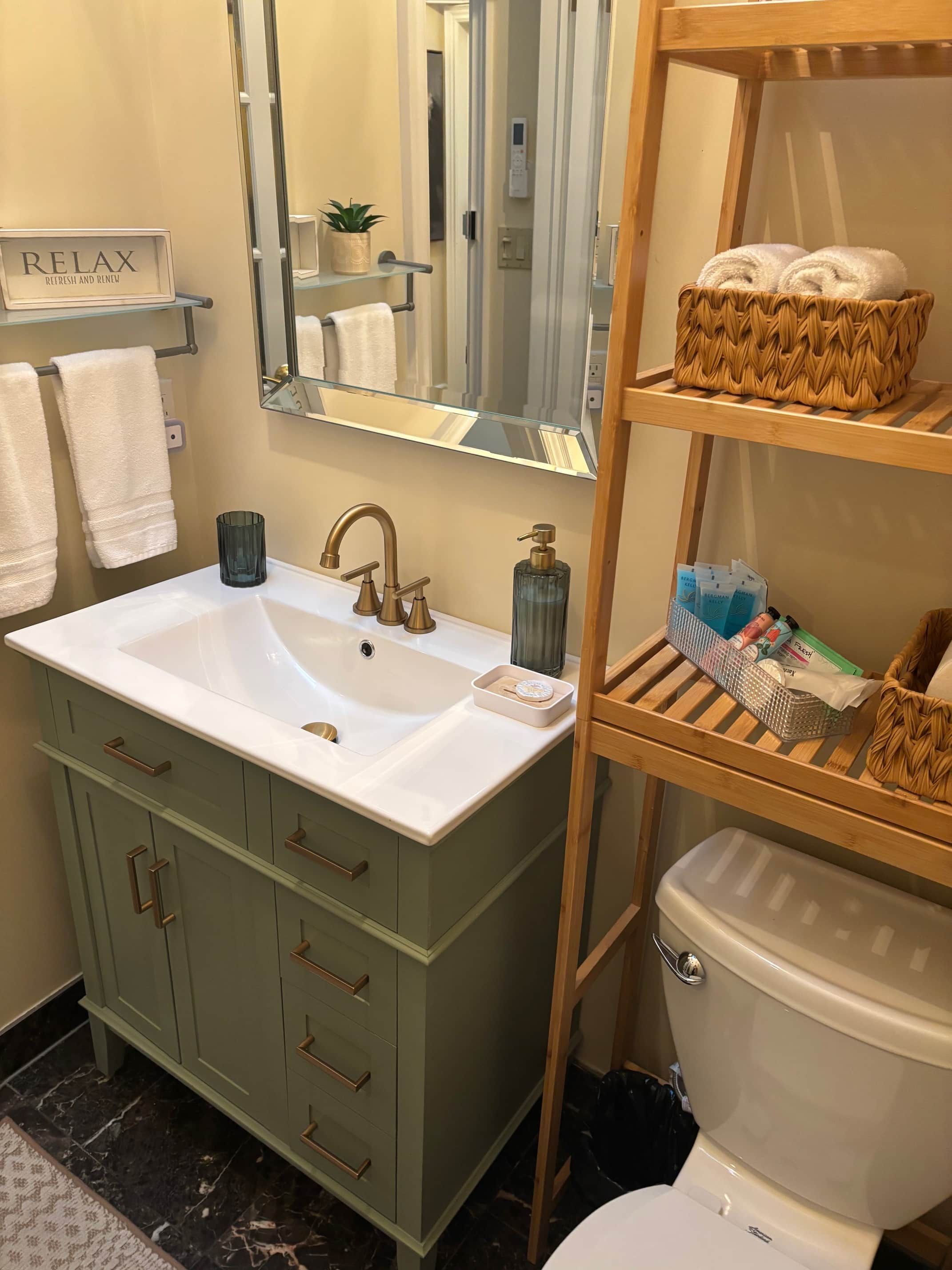 Ein Badezimmer mit einem weißen Waschbecken, einem Spiegel, einem Handtuchhalter, einer Toilette und einem Regal mit Körben.
