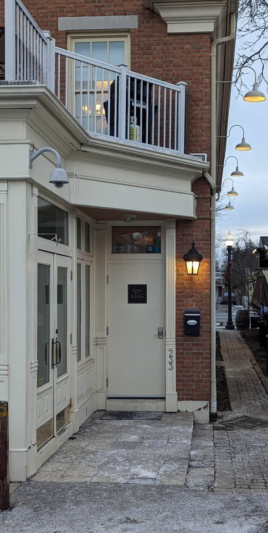 A building with a white facade, a balcony with railings, a door labeled 'SUITES KING,' and a brick wall.