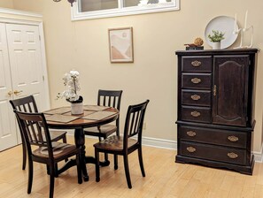 A dining area with a round table, four chairs, a dark wooden cabinet with drawers, a framed picture on the wall, and a window above the table. - The Lilibet Suite - Regal and Royal (Niagara-on-the-Lake)