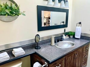 A bathroom with a granite countertop, a round sink, a mirror, and a towel rack. - The Lilibet Suite - Regal and Royal (Niagara-on-the-Lake)