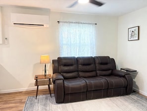 A living room with a leather sofa, a side table with a lamp, a wall-mounted air conditioner, and a ceiling fan.