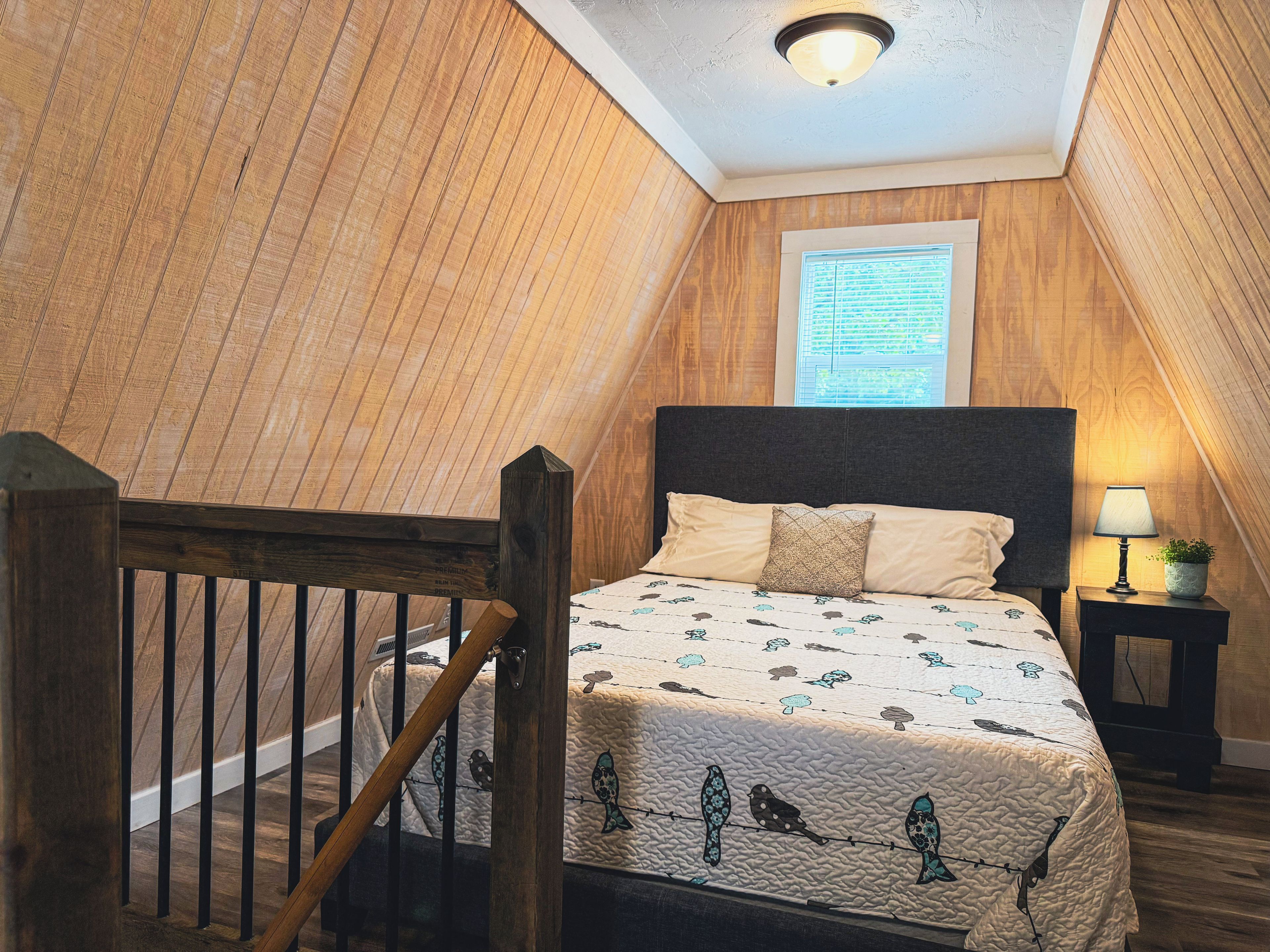 A bedroom with a sloped ceiling, a bed with a patterned quilt, a nightstand with a lamp, and a window with blinds.