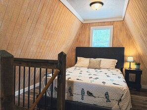 Un dormitorio con techo inclinado, una cama con colcha estampada, una mesita de noche con lámpara y un ventanal con persianas.