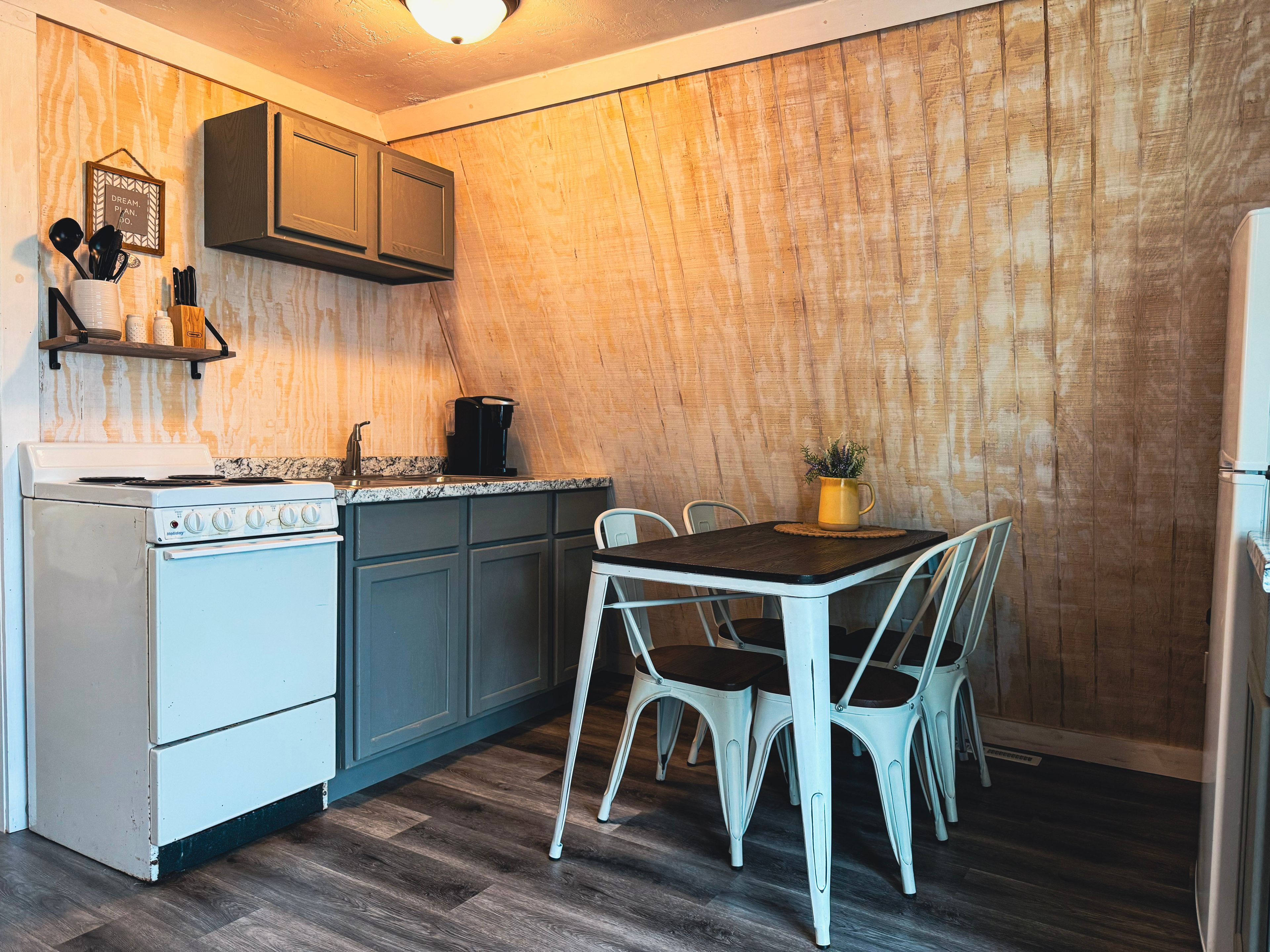 A compact kitchen with a stove, sink, and cabinets, a dining table with chairs, and a wooden wall.