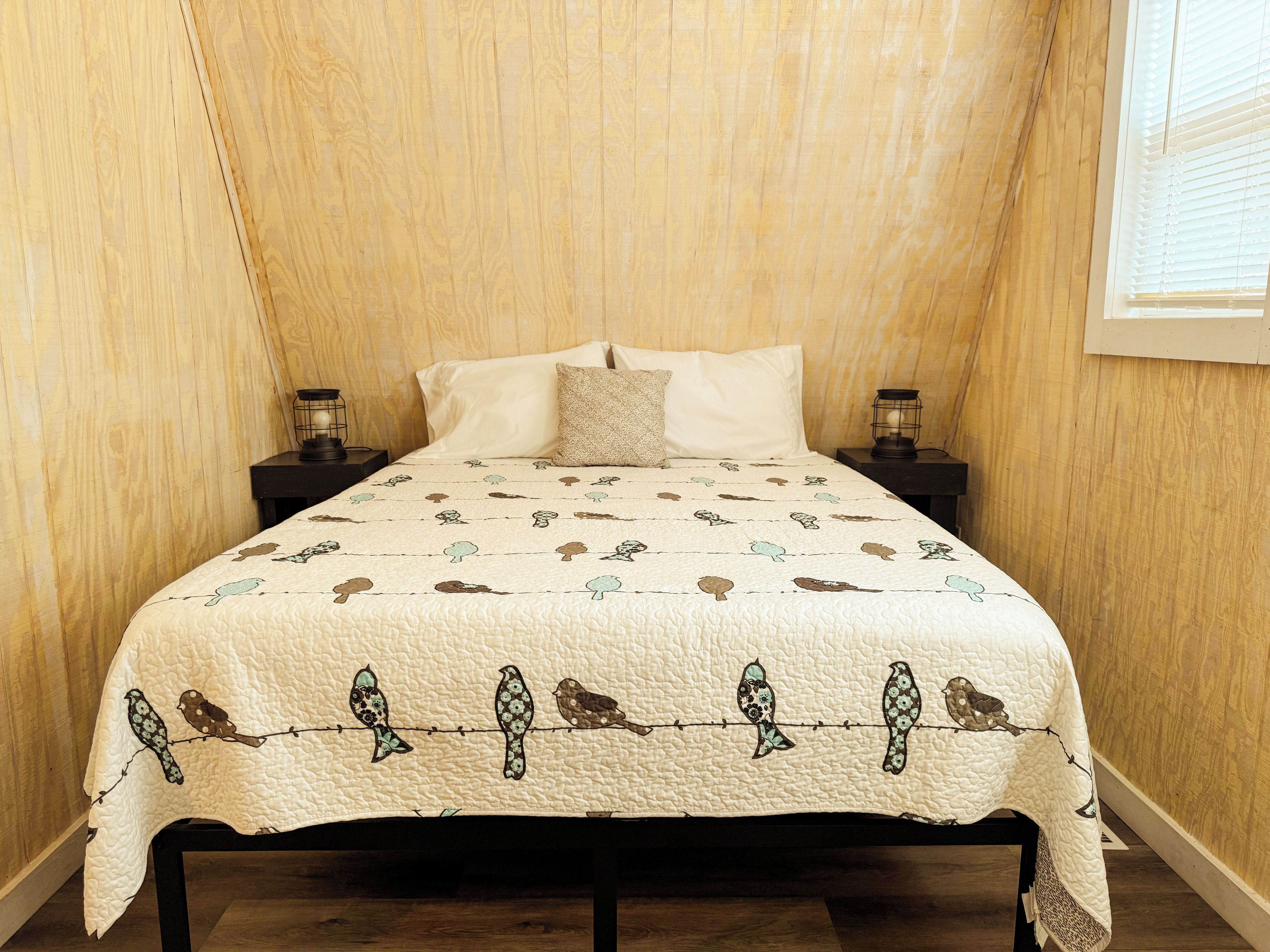 A bed with a quilted cover featuring bird patterns, flanked by two bedside tables with lamps, in a room with wooden walls and a sloped ceiling.