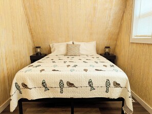 A bed with a quilted cover featuring bird patterns, flanked by two bedside tables with lamps, in a room with wooden walls and a sloped ceiling.