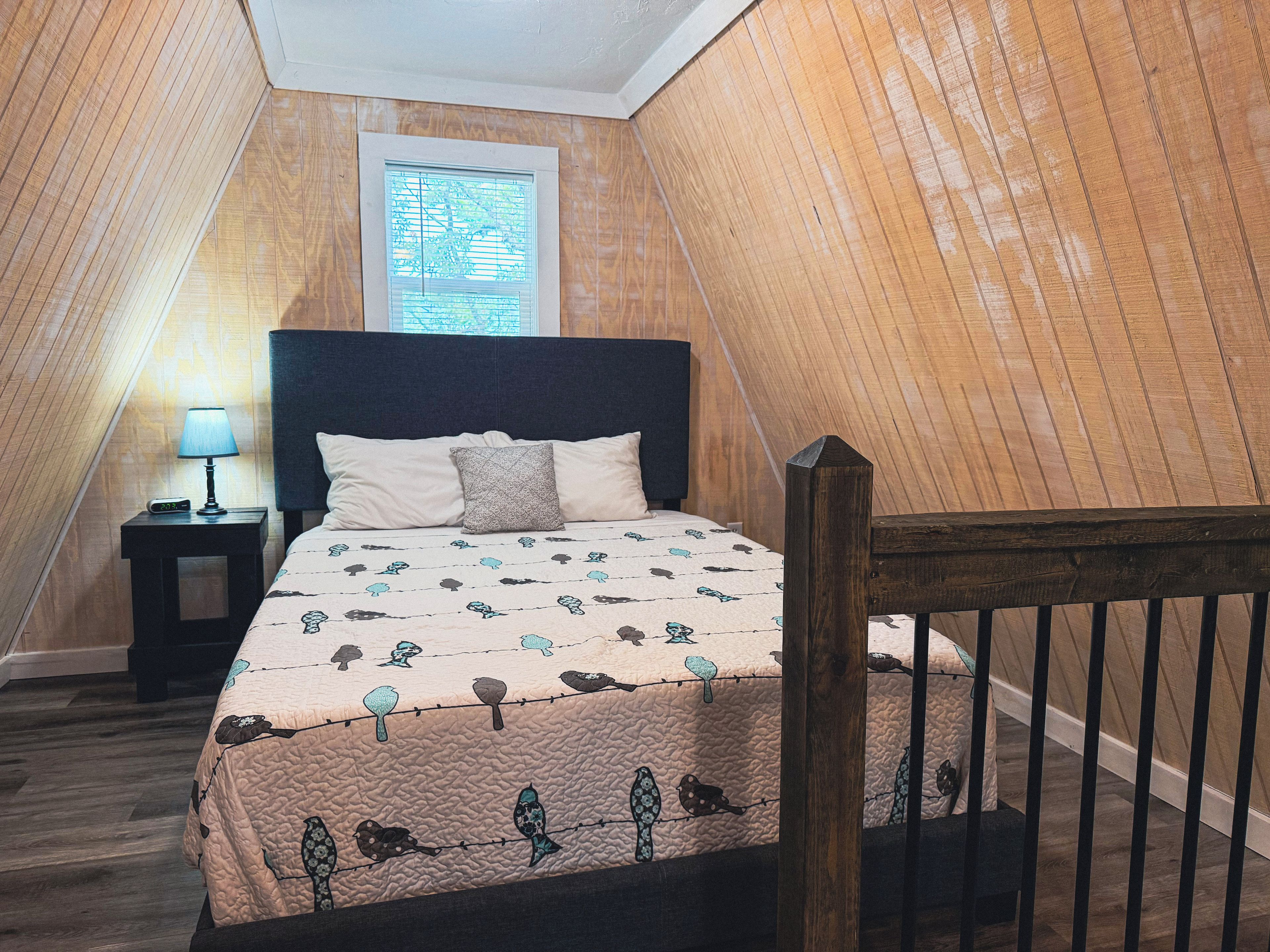 A bedroom with a sloped ceiling, a bed with a patterned bedspread, a nightstand with a lamp, and a window with blinds.