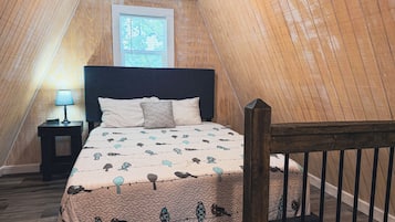 A bedroom with a sloped ceiling, a bed with a patterned bedspread, a nightstand with a lamp, and a window with blinds.