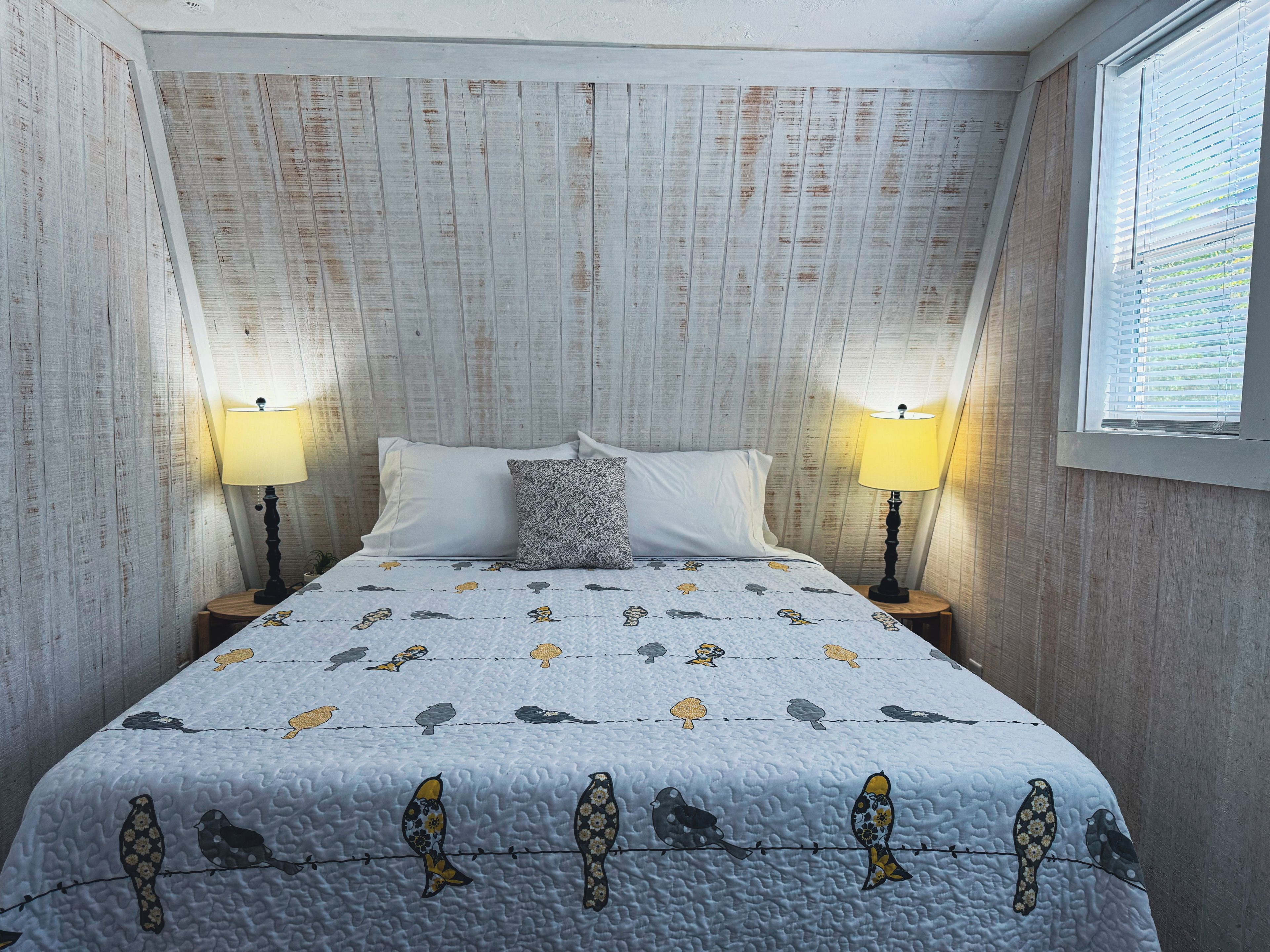 Ein Schlafzimmer mit einem Bett, zwei Nachttischlampen, einem Fenster mit Jalousien und einer gemusterten Tagesdecke.