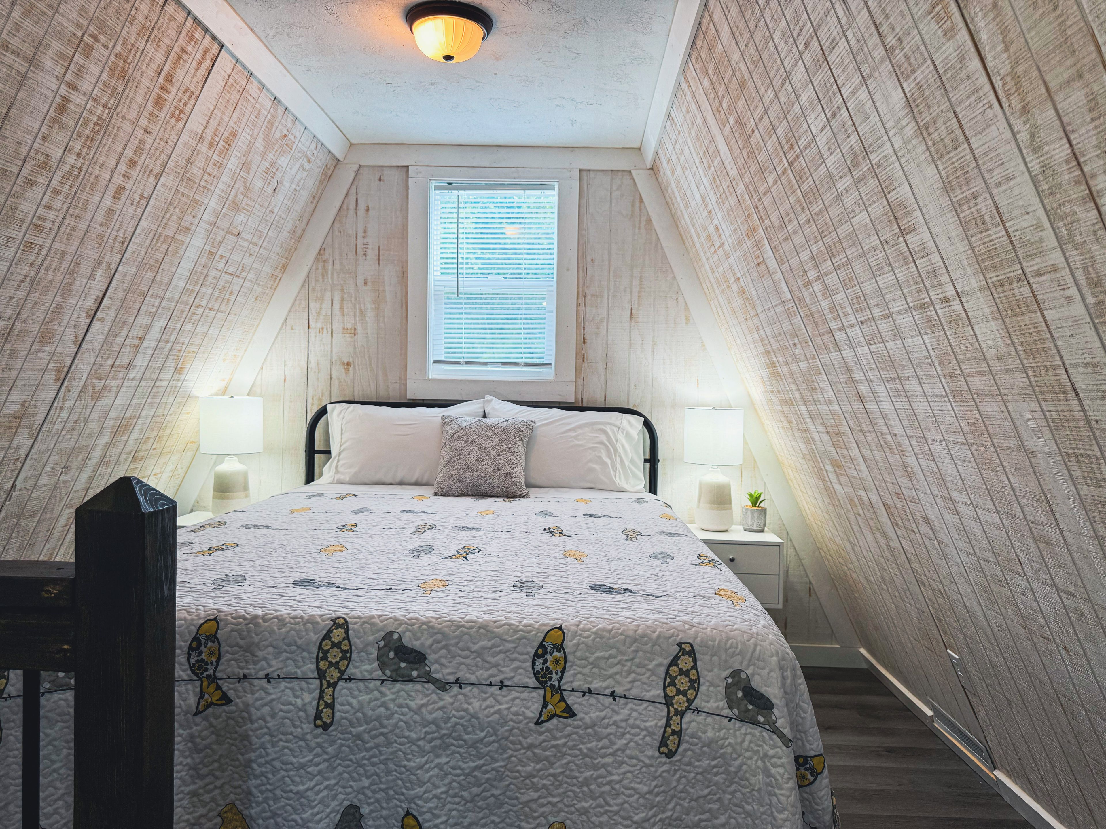 A bedroom with a sloped ceiling, a bed with a patterned quilt, two bedside lamps, and a window with blinds.