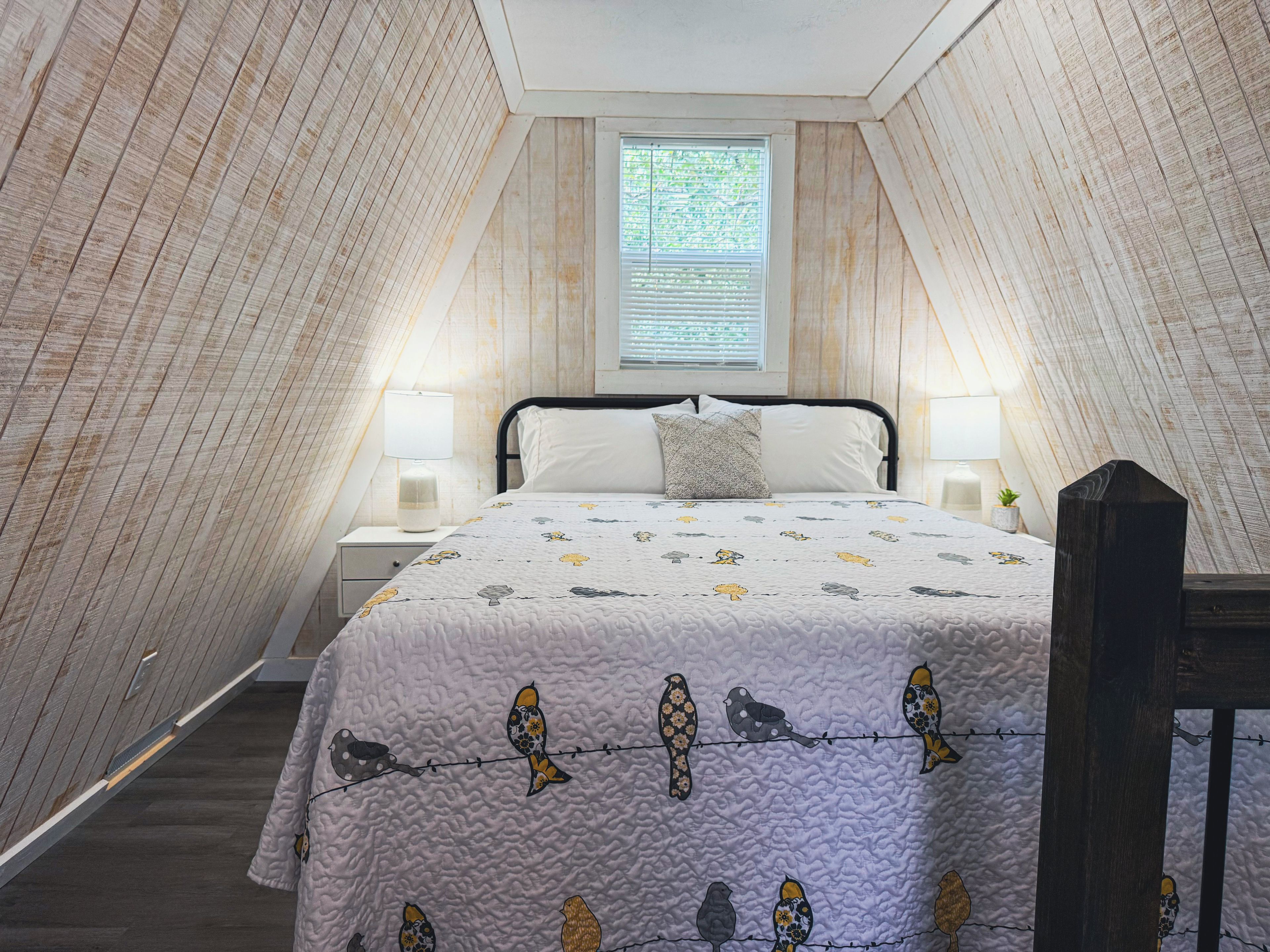 A bedroom with a sloped ceiling, a bed with a patterned duvet, two bedside lamps, and a window with blinds.