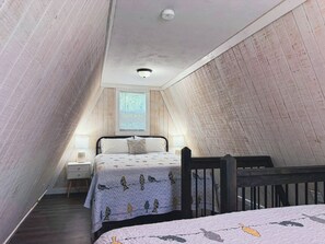 Quarto com teto inclinado, cama com colcha estampada, mesa de cabeceira com abajur e janela com persianas.