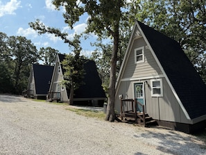 Fileira de pequenas cabanas triangulares com um acesso por caminho de cascalho, cercada por árvores.