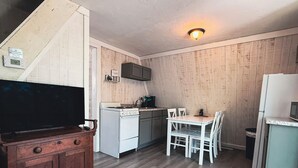 Cocina compacta con estufa, lavamanos y armarios blancos. Se observa una mesa de comedor con sillas y un televisor sobre un mueble de madera.
