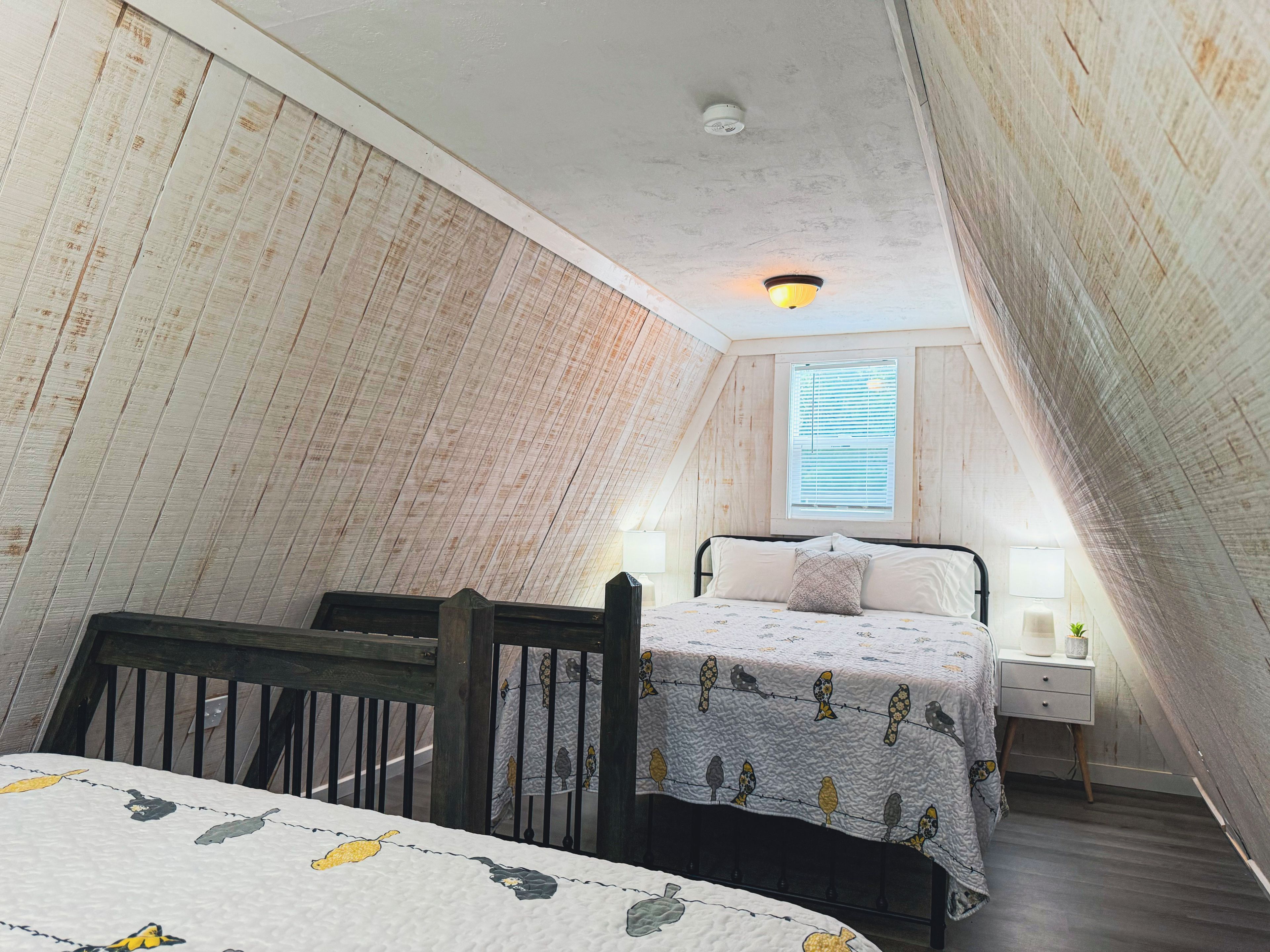 Une chambre avec un plafond en pente, un lit recouvert d’une couvre-lit à motifs, une table de chevet avec une lampe et une fenêtre avec des stores.