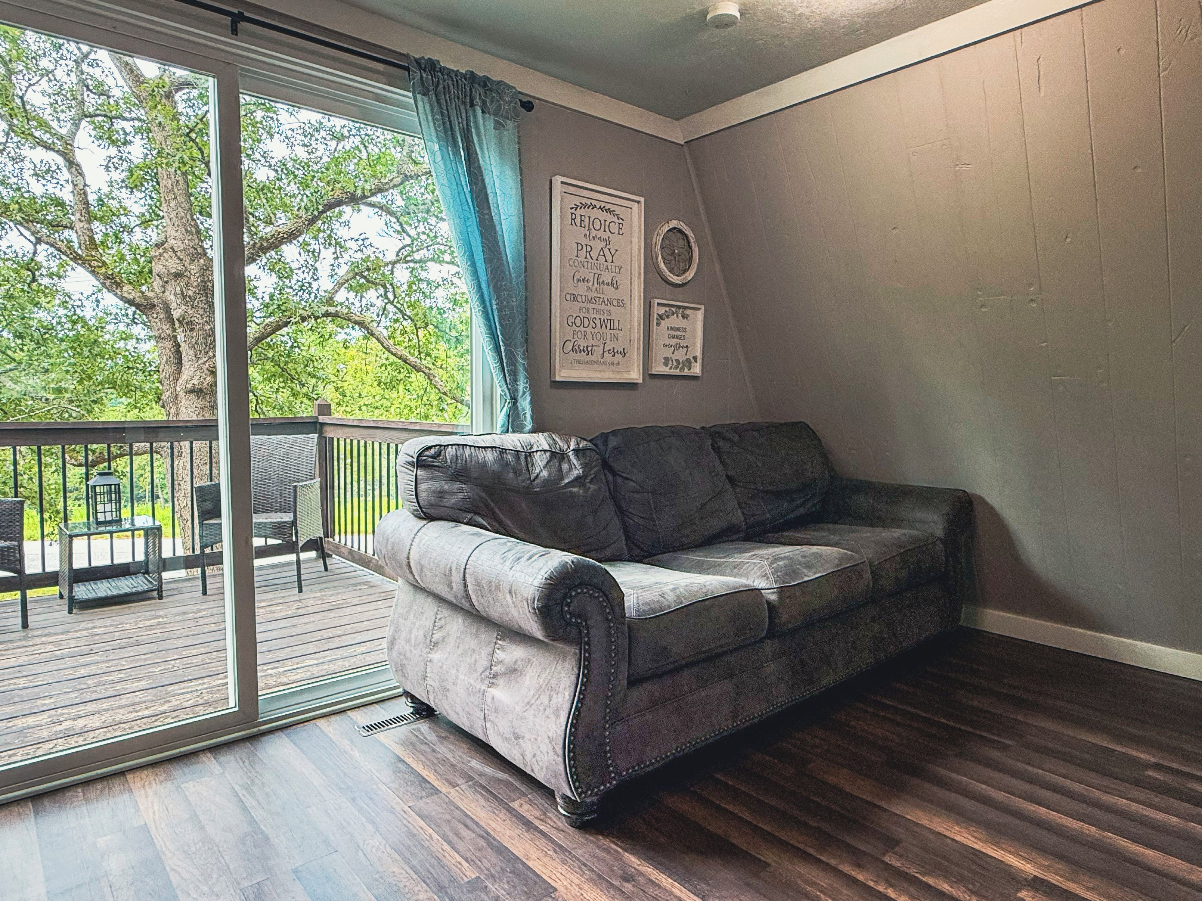 A room with a grey sofa, a sliding glass door leading to a balcony, and a wall with framed inspirational quotes.