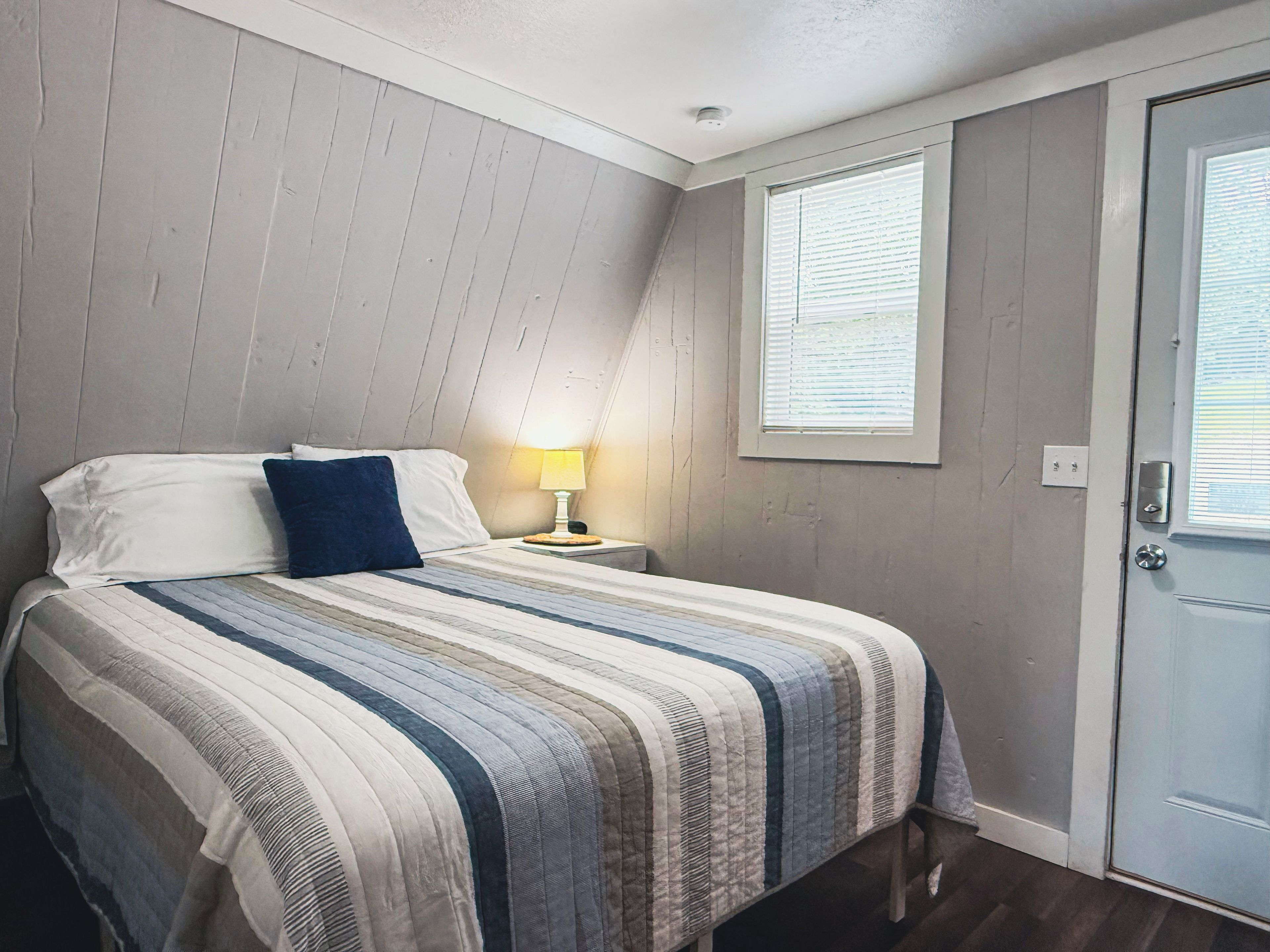 A bedroom with a bed, a nightstand, a lamp, a window, and a door.