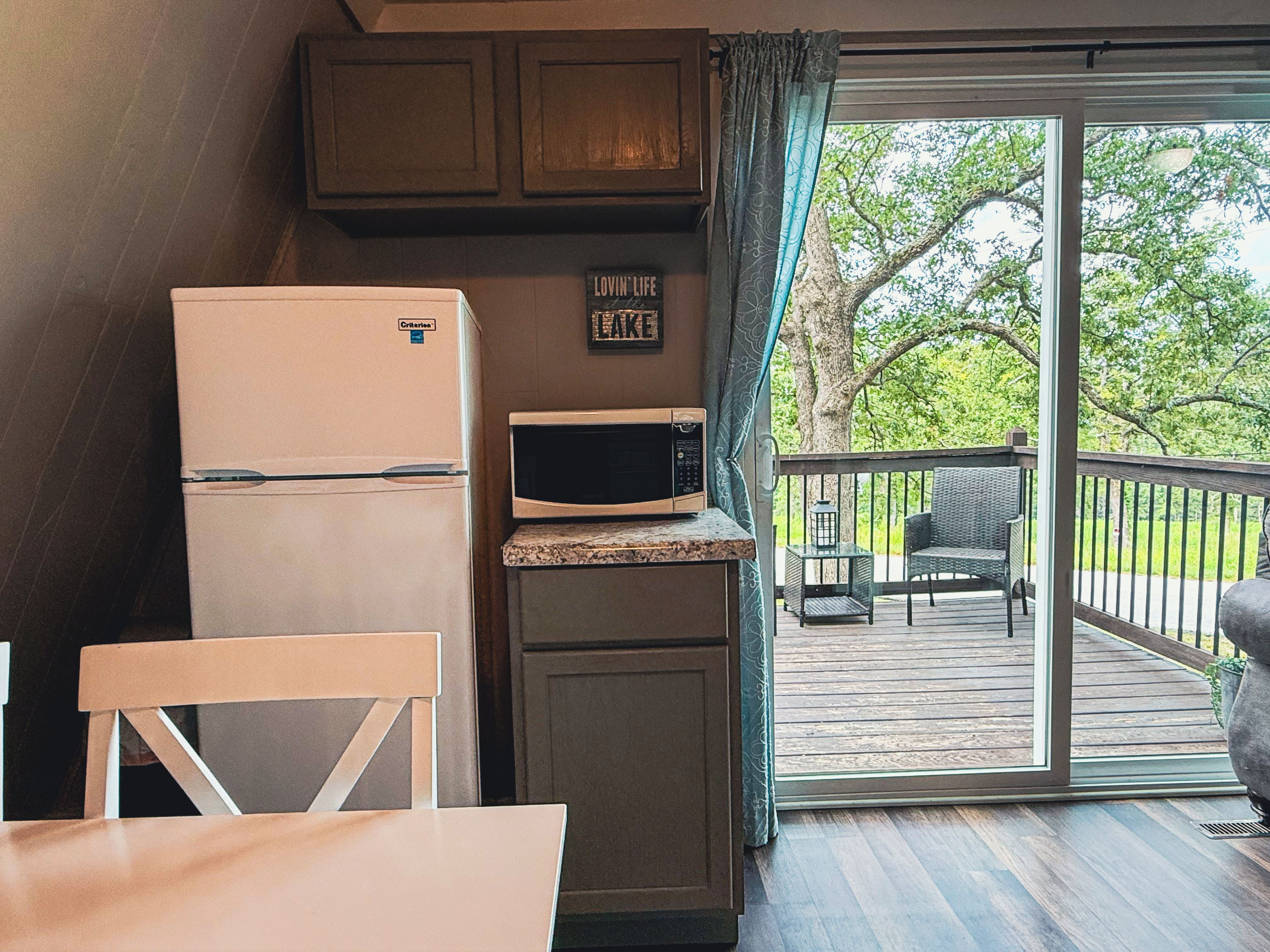 Una kitchenette compacta con refrigerador, microondas y un mueble pequeño. Hay una zona de comedor con una mesa y sillas, y un balcón con vista a árboles.