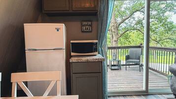 Una kitchenette compacta con refrigerador, microondas y un mueble pequeño. Hay una zona de comedor con una mesa y sillas, y un balcón con vista a árboles.