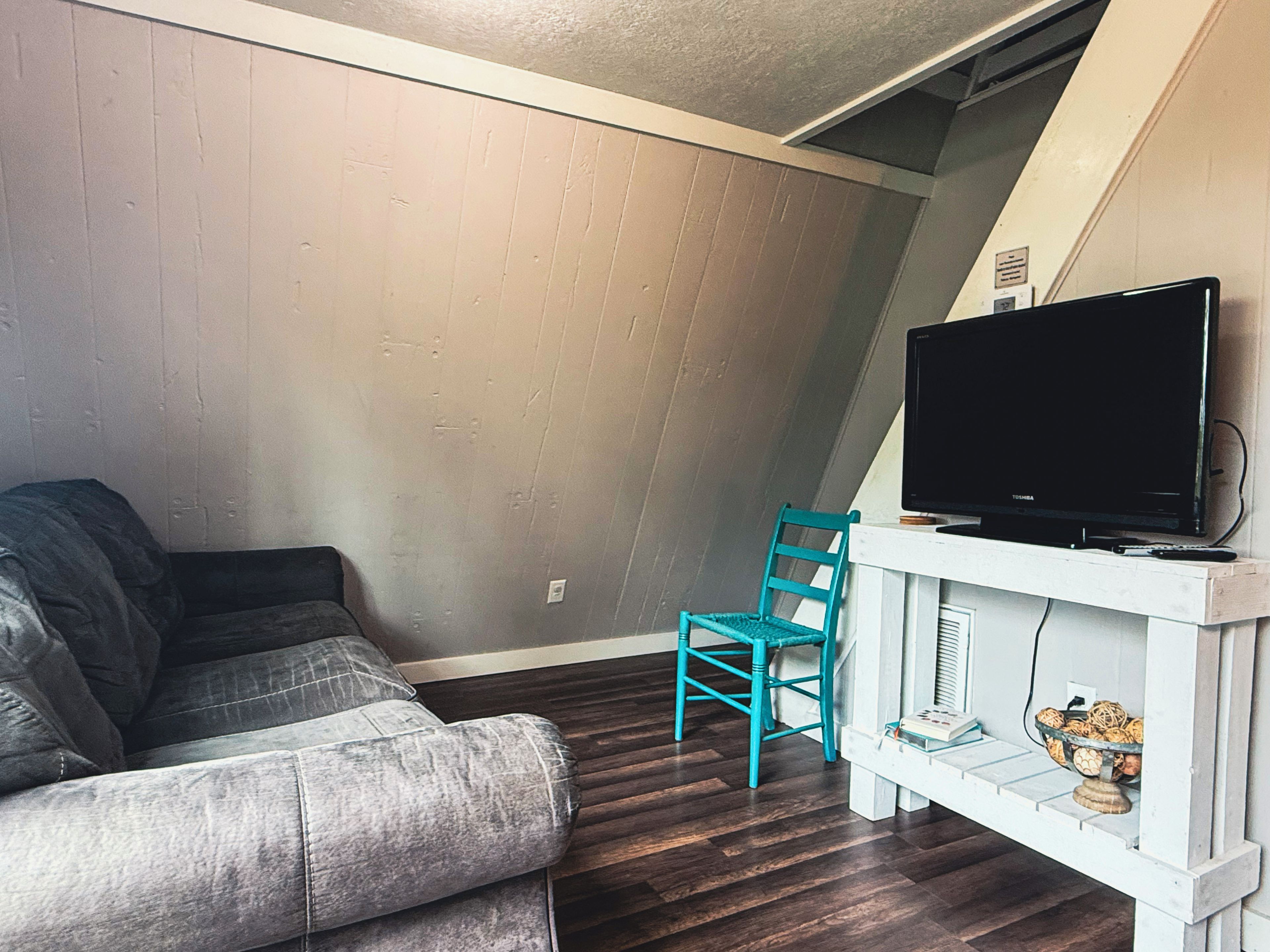 Una sala de estar con un sofá gris, un sillón azul turquesa, un televisor de pantalla plana sobre un estante blanco y un estante pequeño blanco con libros y un cuenco.