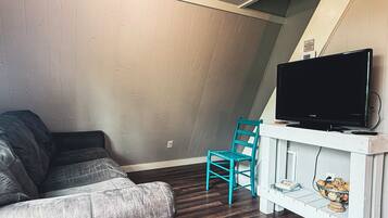 Una sala de estar con un sofá gris, un sillón azul turquesa, un televisor de pantalla plana sobre un estante blanco y un estante pequeño blanco con libros y un cuenco.