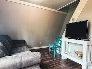 Een woonkamer met een grijze bank, een blauwgroene fauteuil, een flatscreen televisie op een witte standaard, en een klein wit plankje met boeken en een schaal.