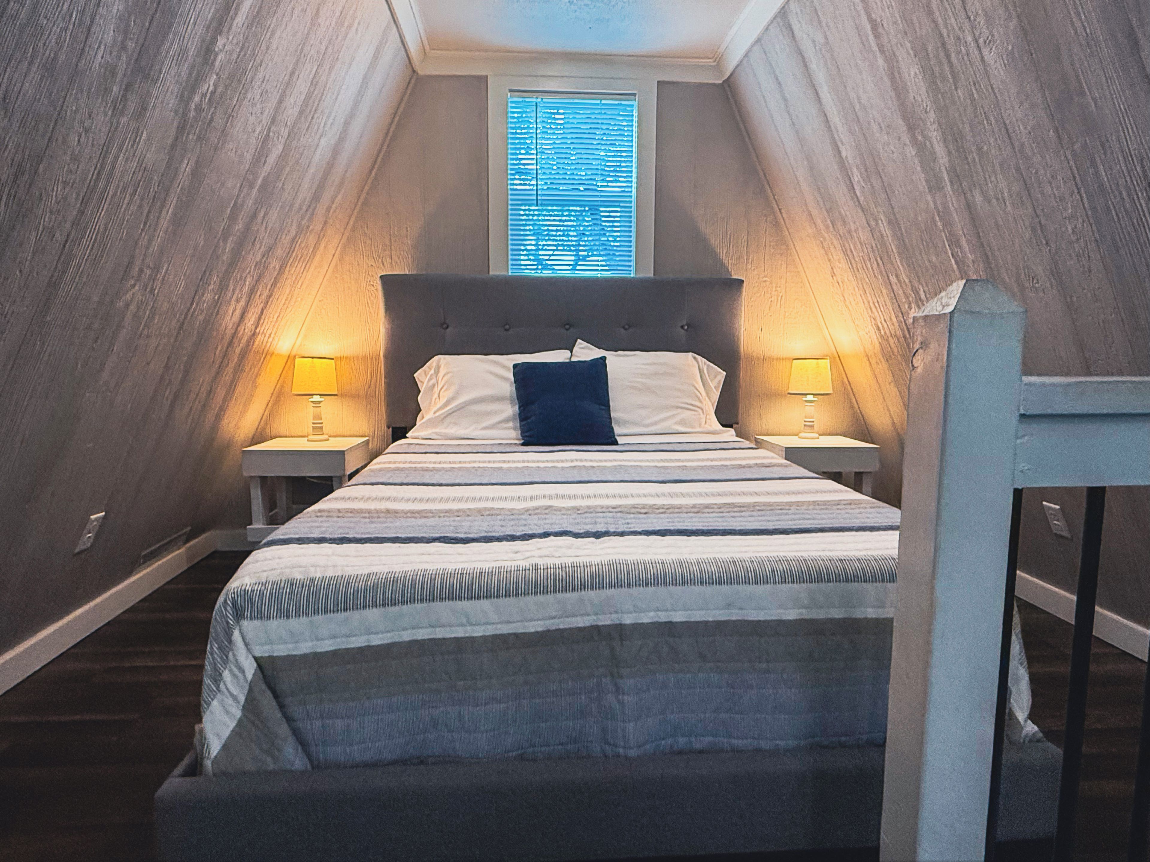 A bedroom with a sloped ceiling, a bed with a headboard, two bedside tables with lamps, and a window with blinds.