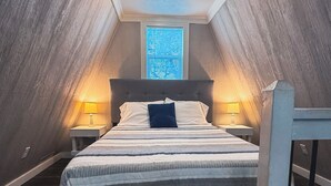 Een slaapkamer met een schuine kap, een bed met een gestreept sprei, twee bedlampen en een raam met blinds.