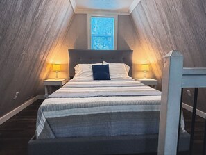 Un dormitorio con techo inclinado, una cama con colcha a rayas, dos lámparas de noche y una ventana con persianas.