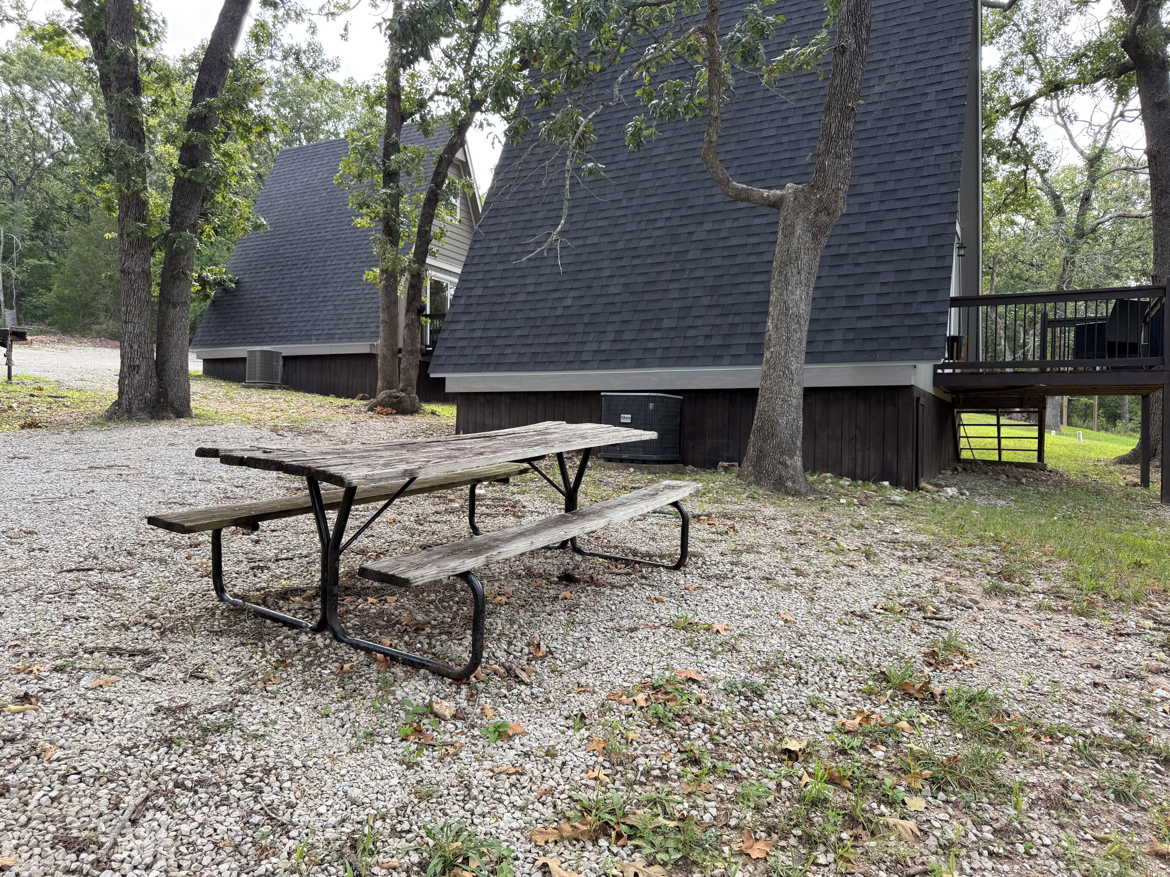 A picnic table with benches in a gravel area with trees and a building in the background.