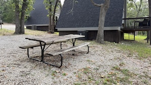 Een picknicktafel met banken in een grindgebied, omringd door bomen en een gebouw met een donker dak.
