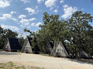 Fileira de cabanas pequenas com telhados triangulares, um caminho de cascalho e árvores ao fundo.