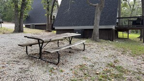 Una mesa de picnic con bancos en un área de grava, rodeada de árboles y un edificio con techo oscuro.