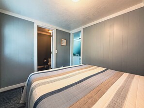 Un dormitorio con cama, una puerta que da al baño y una lámpara fijada a la pared.