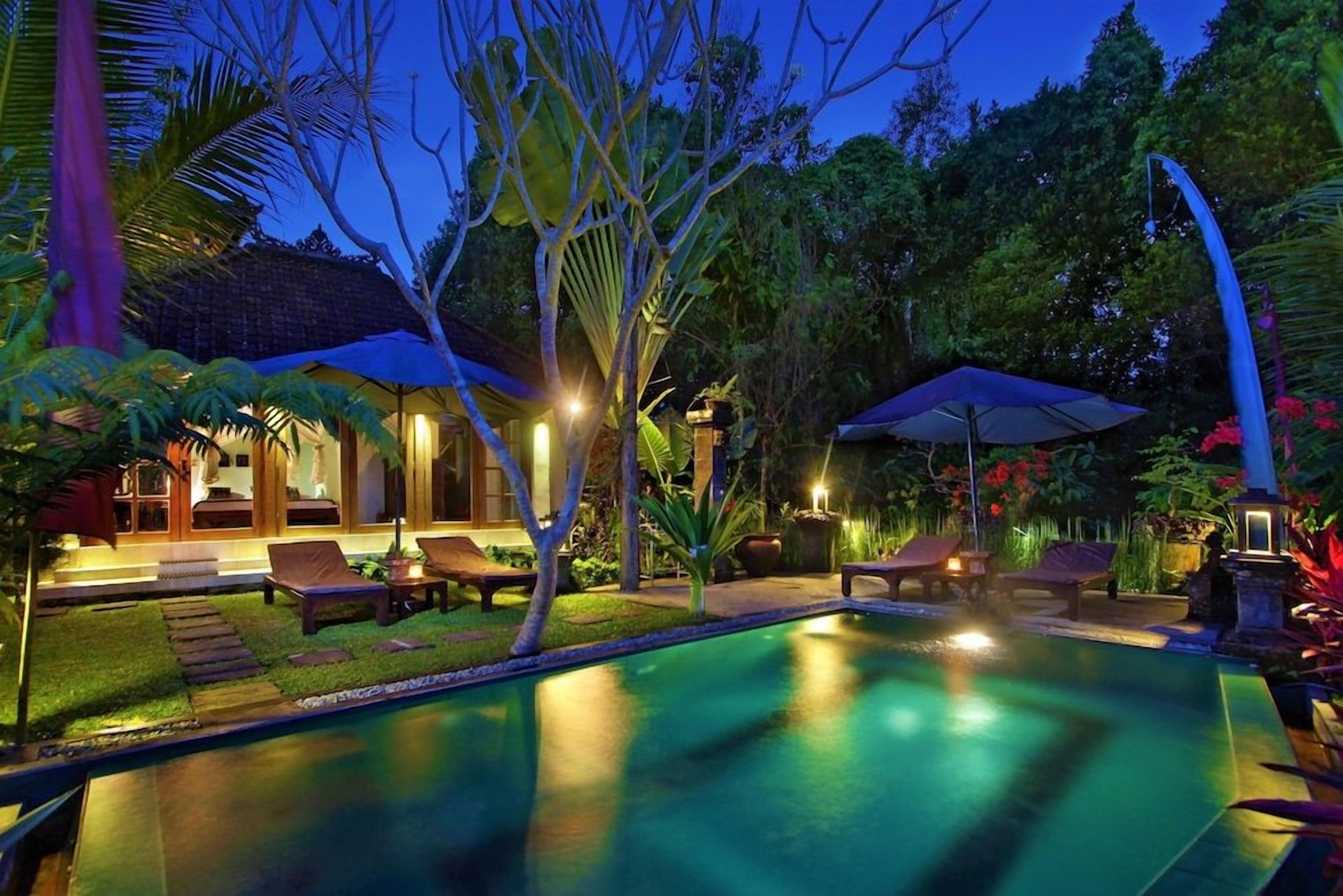 A swimming pool with lounge chairs and umbrellas, surrounded by tropical vegetation and a building with a tiled roof.