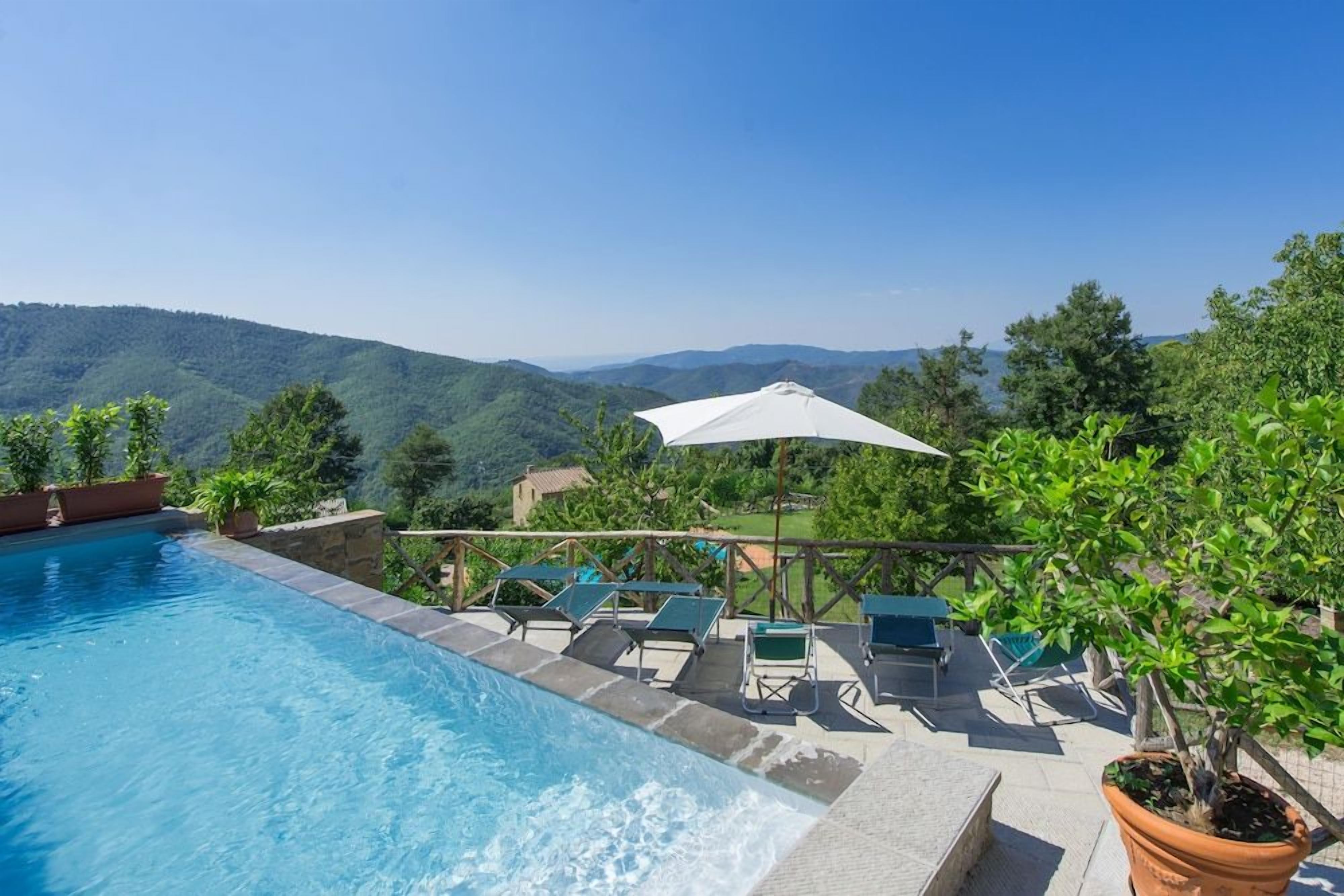 A swimming pool with a view of hills and a pergola with umbrellas and lounge chairs.