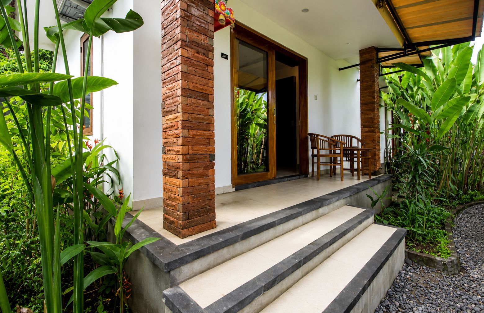 A covered outdoor area with steps leading to a door, surrounded by lush greenery.
