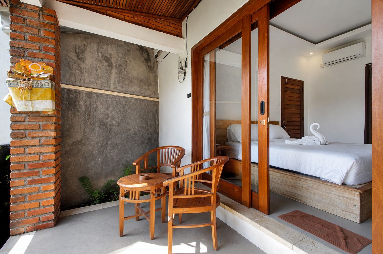 A room with a bed, a wooden table with chairs, a brick wall, and a sliding glass door leading to a balcony.